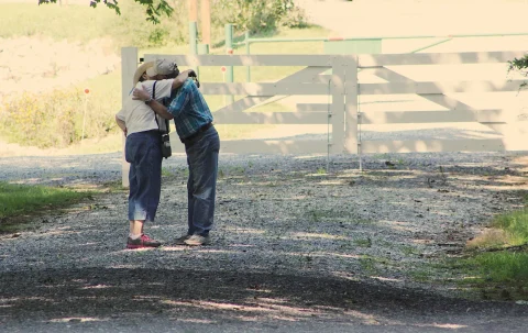 An elderly couple embrace in front of a large white farm gate
