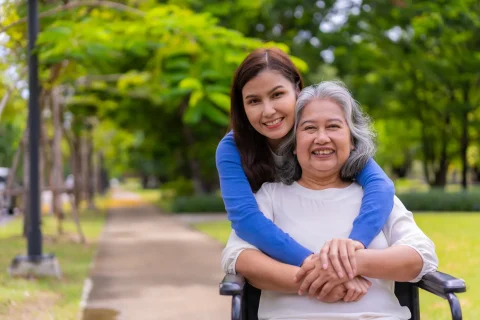 A young woman hugs an older woman in a wheelchair from behind while holding hands.