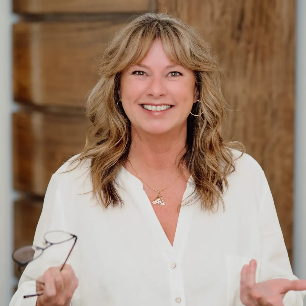 Kimberley smiling in a white blouse hands in front, right hand holding glasses.
